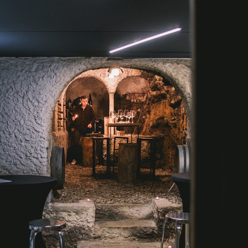 L'interno di una suggestiva cantina con pareti di pietra grezza e un arco illuminato. Un uomo in camicia scura si trova dietro a una postazione degustazione creata con tronchi di legno e botti, dove sono disposti dei calici di vino, in un'atmosfera intima e accogliente. | © Filippo Frizzera, 2025