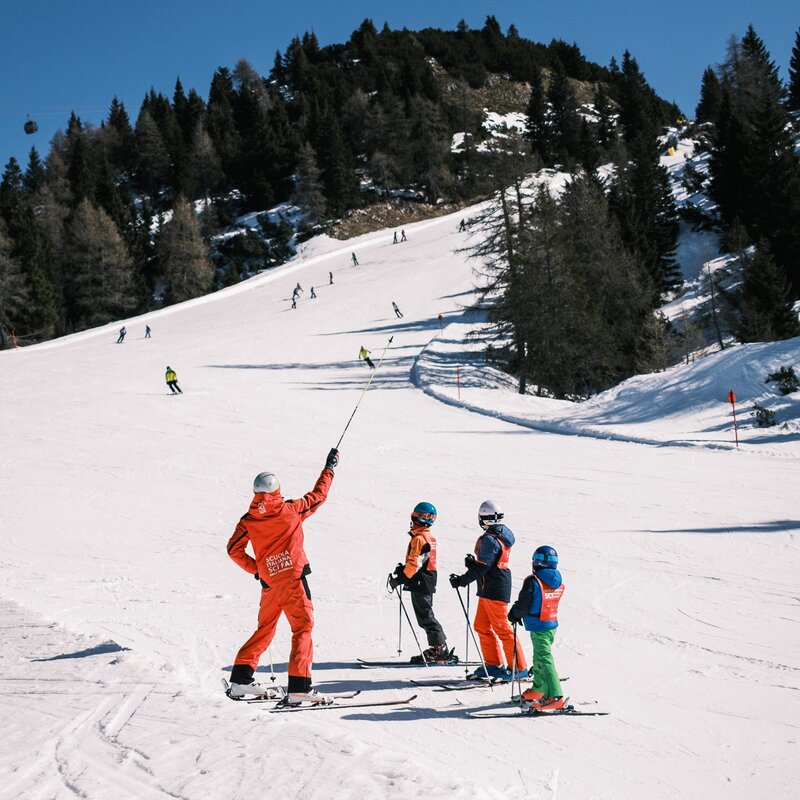 Imparare a sciare in Paganella | © Filippo Frizzera, 2025 Su un'ampia pista da sci della Paganella, un maestro di sci in divisa rossa, visto di schiena, dà istruzioni a tre bambini. Il maestro indica con un bastoncino verso la cima della pista, circondata da un bosco di conifere. | © Filippo Frizzera, 2025