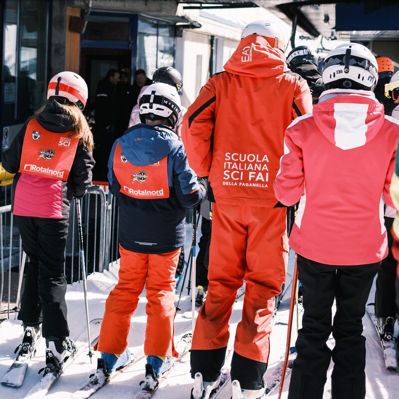 Imparare a sciare in Paganella | © Filippo Frizzera, 2025 Un gruppo di bambini, con sci e caschetti, è in fila sulla neve insieme a un maestro della "Scuola Italiana Sci Fai della Paganella". Visti di schiena, sono in attesa di prendere un impianto di risalita. | © Filippo Frizzera, 2025