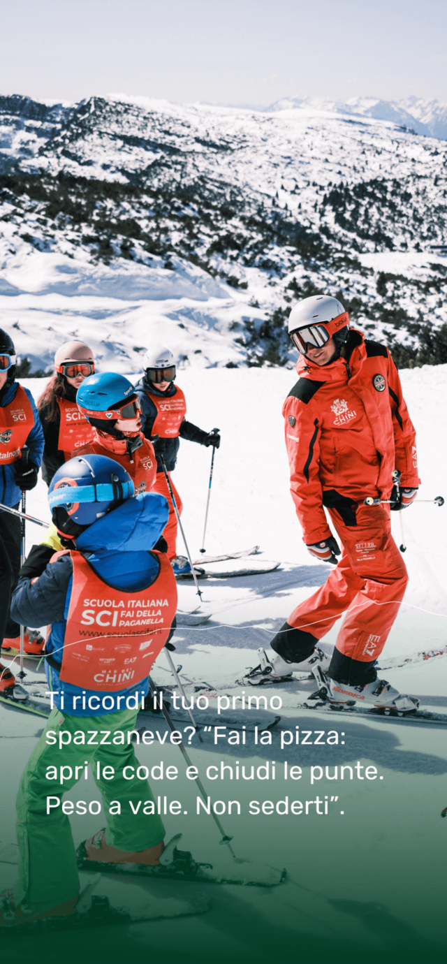 Un maestro di sci della Paganella, in divisa rossa, mostra la tecnica di sci a un gruppo di bambini durante una lezione di sci. Sullo sfondo, si apre un ampio panorama di piste innevate.  Testo in sovrimpressione: Ti ricordi il tuo primo spazzaneve? “Fai la pizza: apri le code e chiudi le punte. Peso a valle. Non sederti”. | © Filippo Frizzera, 2025
