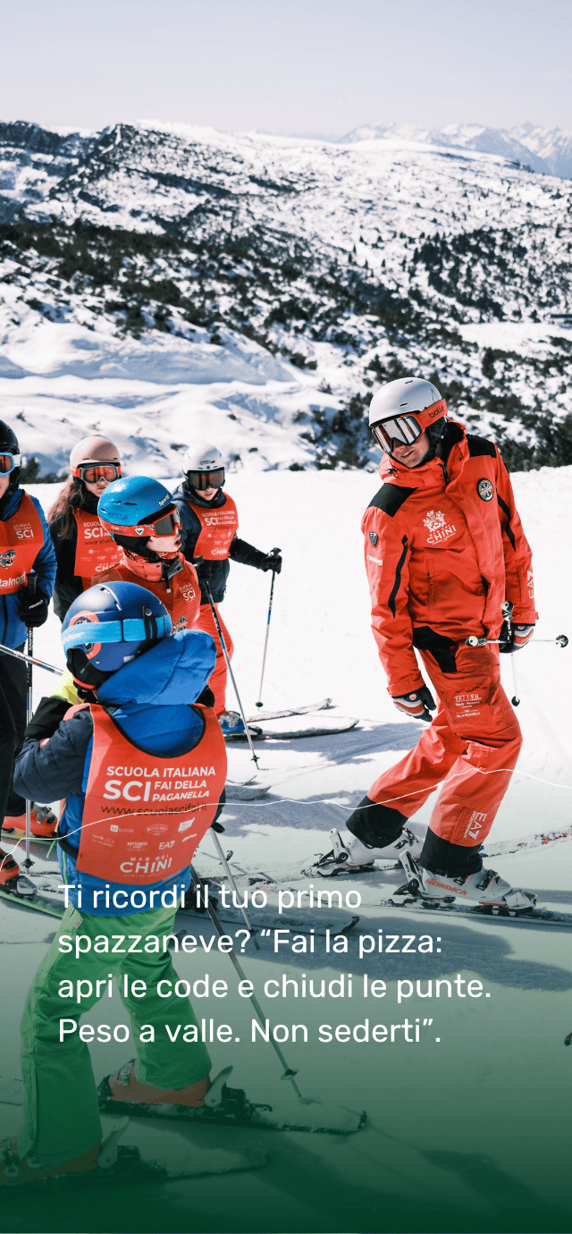 Un maestro di sci della Paganella, in divisa rossa, mostra la tecnica di sci a un gruppo di bambini durante una lezione di sci. Sullo sfondo, si apre un ampio panorama di piste innevate.  Testo in sovrimpressione: Ti ricordi il tuo primo spazzaneve? “Fai la pizza: apri le code e chiudi le punte. Peso a valle. Non sederti”. | © Filippo Frizzera, 2025