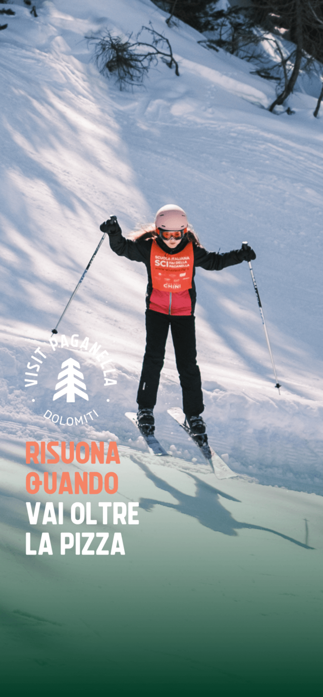 Una bambina scia con sicurezza su una pista innevata della Paganella, indossando casco, occhiali da sci e una pettorina arancione della scuola di sci. Il testo in sovrimpressione dice "RISUONA QUANDO VAI OLTRE LA PIZZA". | © Filippo Frizzera, 2025