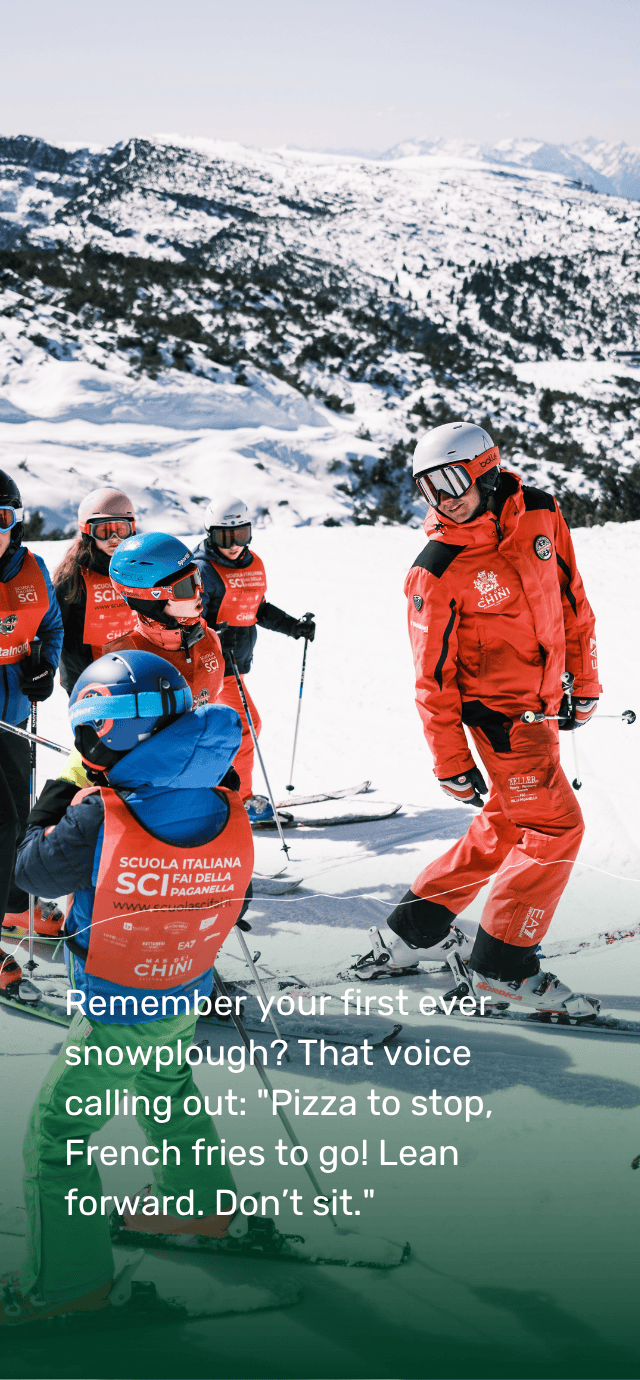 A ski instructor from Paganella, dressed in a red uniform, demonstrates skiing technique to a group of children during a ski lesson. In the background, there is a wide panorama of snow-covered slopes.  Overlay text: Remember your first ever snowplough? That voice calling out: "Pizza to stop, French fries to go! Lean forward. Don’t sit." | © Filippo Frizzera, 2025