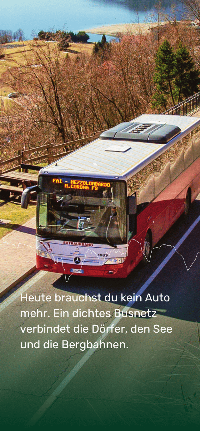 Eine Aufnahme von schräg oben auf einen roten und grauen Überlandbus, der auf einer asphaltierten Straße fährt. Die Leuchtanzeige auf der Vorderseite des Busses zeigt "FAI - MEZZOLOMBARDO M.CORONA FS". Im Hintergrund sind grasbewachsene Hügel, kahle Bäume und das Ufer eines Sees zu sehen. Etwa auf halber Höhe verläuft eine dünne, unregelmäßige weiße Linie, die an die stilisierte Silhouette von Bergen erinnert. Im unteren Bereich steht auf einem dunkelgrünen Farbverlauf der folgende Text in Weiß: "Heute brauchst du kein Auto mehr. Ein dichtes Busnetz verbindet die Dörfer, den See und die Bergbahnen." | © Filippo Frizzzera, 2019