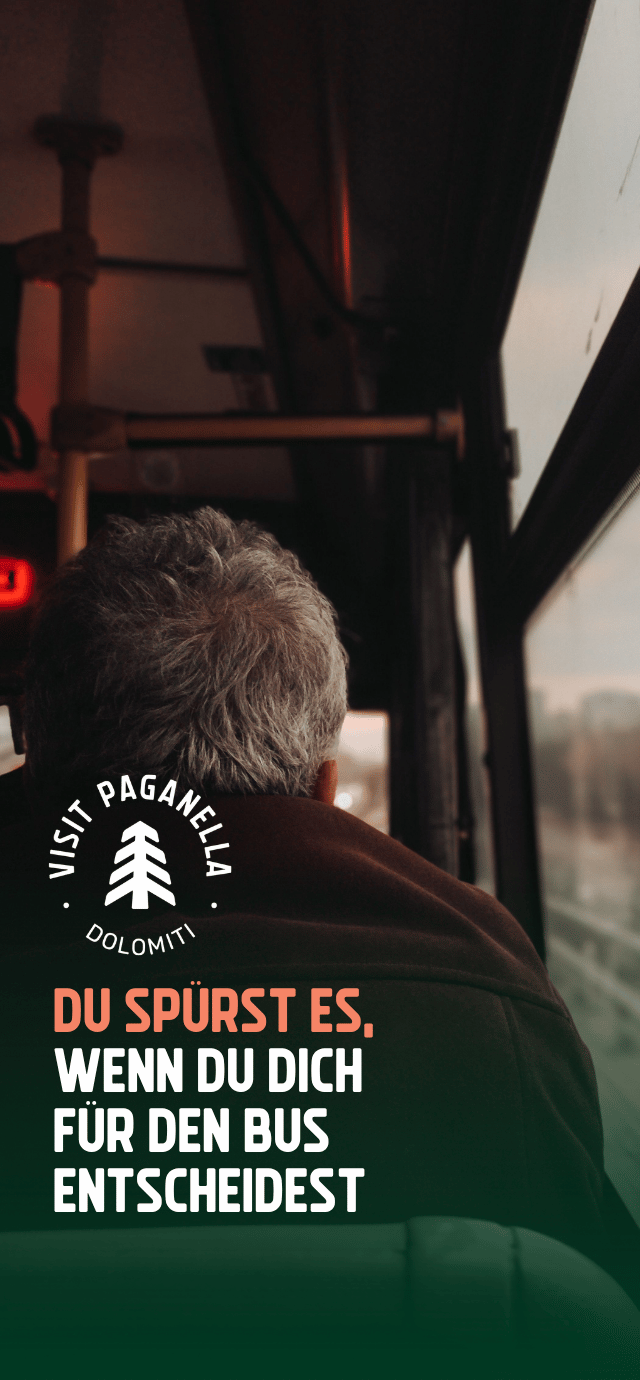 Die Ansicht aus dem Inneren eines Busses, die über die Schulter eines Fahrgastes mit grauem Haar blickt, der aus dem Fenster schaut. Die Atmosphäre ist von warmem Licht und Schatten geprägt. Unten links befindet sich auf einem dunkelgrünen Farbverlauf ein kreisförmiges weißes Logo mit einer stilisierten Tanne in der Mitte und den Wörtern "VISIT PAGANELLA" und "DOLOMITI". Unter dem Logo steht in Großbuchstaben der folgende Text: "DU SPÜRST ES," (in Pfirsichfarbe) "WENN DU DICH FÜR DEN BUS ENTSCHEIDEST" (in Weiß) | © Tuğba Özsoy