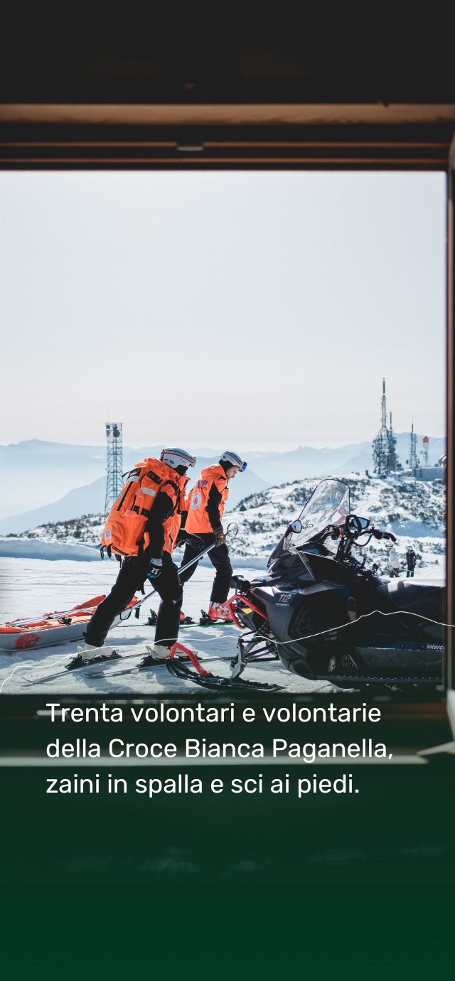 Vista dall'interno verso l'esterno di due soccorritori in divisa arancione e nera che camminano sulla neve accanto a una motoslitta e a una barella arancione. Sullo sfondo, montagne e alte antenne per le telecomunicazioni sotto un cielo sereno. In basso, il testo recita: "Trenta volontari e volontarie della Croce Bianca Paganella, zaini in spalla e sci ai piedi." | © Filippo Frizzera, 2026