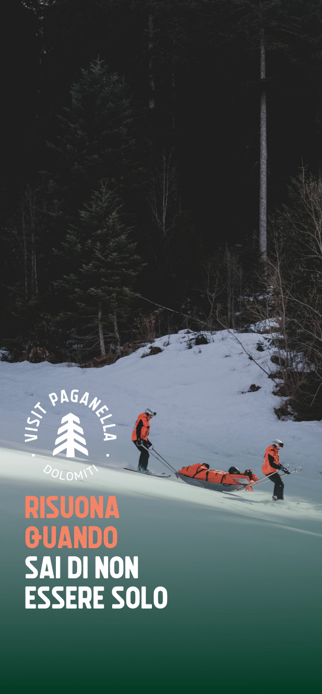 Due soccorritori con giacca arancione scendono con gli sci su un pendio innevato, guidando una barella arancione di salvataggio. Sullo sfondo si erge un fitto e scuro bosco di conifere. In basso a sinistra, sotto il logo Visit Paganella, campeggia la scritta "RISUONA QUANDO SAI DI NON ESSERE SOLO". | © Filippo Frizzera, 2026