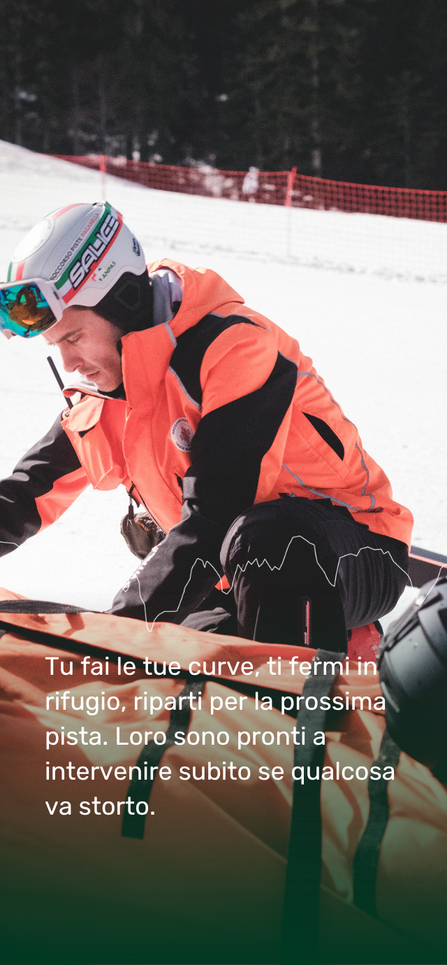 Primo piano di un soccorritore accovacciato sulla neve accanto a una barella arancione da neve. L'uomo indossa una spessa giacca arancione e nera e un casco protettivo. Il testo bianco in sovrimpressione recita: "Tu fai le tue curve, ti fermi in rifugio, riparti per la prossima pista. Loro sono pronti a intervenire subito se qualcosa va storto." | © Filippo Frizzera, 2026