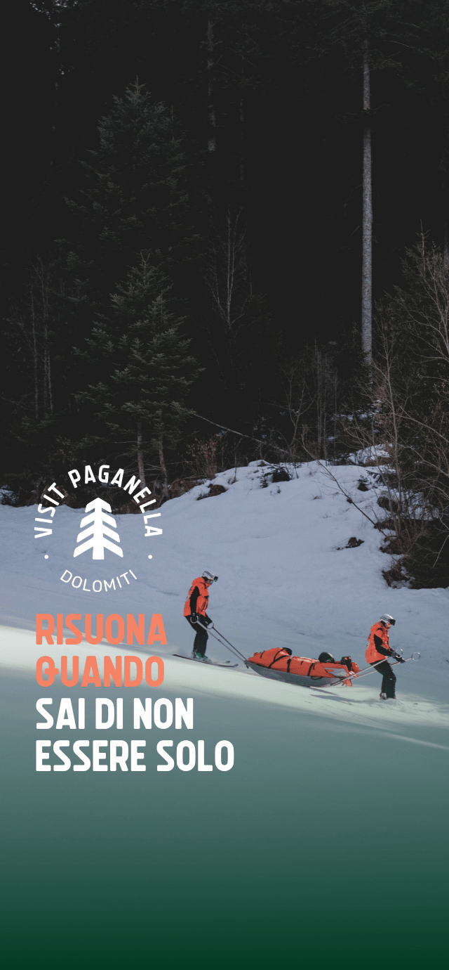 Due soccorritori con giacca arancione scendono con gli sci su un pendio innevato, guidando una barella arancione di salvataggio. Sullo sfondo si erge un fitto e scuro bosco di conifere. In basso a sinistra, sotto il logo Visit Paganella, campeggia la scritta "RISUONA QUANDO SAI DI NON ESSERE SOLO". | © Filippo Frizzera, 2026