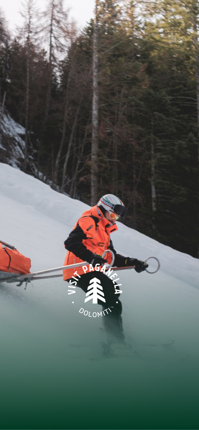 Un soccorritore con giacca arancione, casco e occhiali da sci scende su una pista innevata trainando una barella di salvataggio arancione. Sullo sfondo, un fitto bosco di conifere. Al centro dell'immagine, in sovrimpressione su una sfumatura verde scuro, è presente il logo circolare bianco "VISIT PAGANELLA". | © Filippo Frizzera, 2026