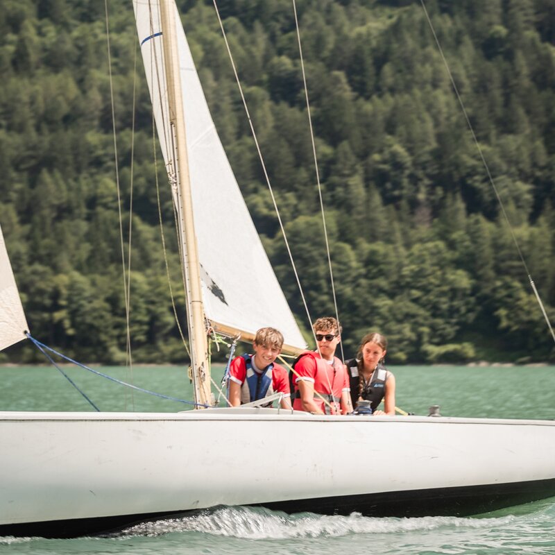 Due ragazzi e una ragazza in navigazione su una barca a vela solcano le acque del lago di Molveno, concentrati e affiatati. Le vele catturano il vento, mentre sullo sfondo la fitta vegetazione della montagna crea un contrasto potente con il bianco dello scafo. | © Filippo Frizzera, 2024