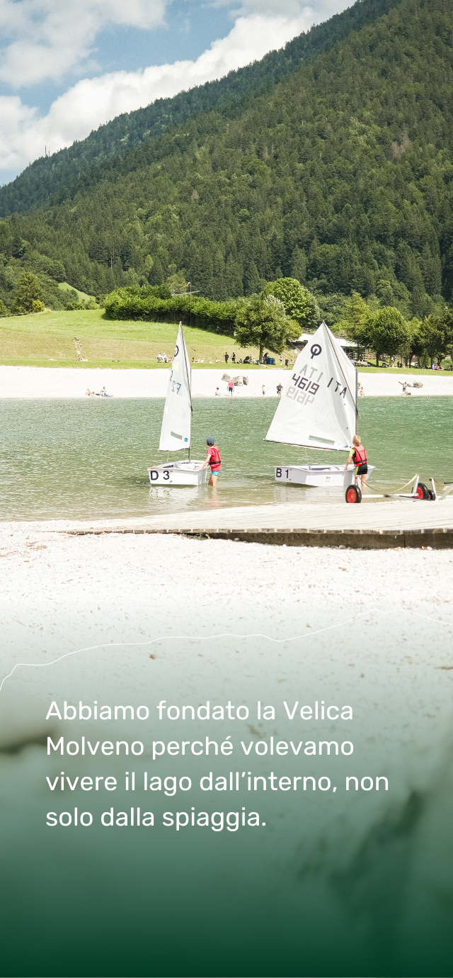 Due bambini con il giubbotto salvagente rosso stanno spingendo in acqua due piccole barche a vela bianche, pronte a salpare sul lago di Molveno. Le vele sono già issate, segnate con numeri e scritte. L’acqua è limpida e tranquilla, incorniciata da fitti boschi verdi che ricoprono le montagne tutt’intorno. In basso, una scritta racconta: “Abbiamo fondato la Velica Molveno perché volevamo vivere il lago dall’interno, non solo dalla spiaggia | © Filippo Frizzera, 2024