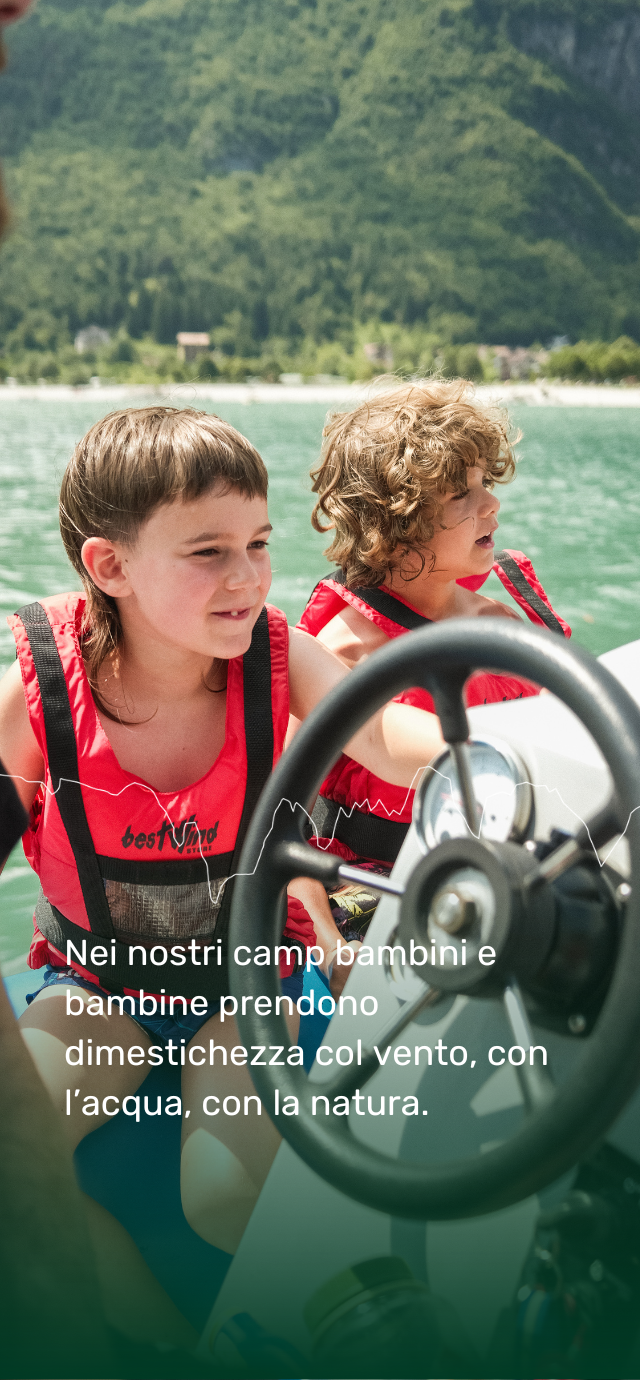 Due bambini con indosso giubbotti salvagente rossi sono seduti su una barca a vela, concentrati e sorridenti. In primo piano si vede il timone. Sullo sfondo, il lago verde è circondato da montagne boscose. In basso, la foto include un testo: “Nei nostri camp bambini e bambine prendono dimestichezza col vento, con l’acqua, con la natura.” | © Filippo Frizzera, 2024