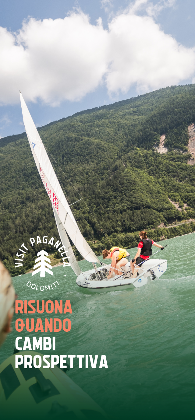 Due persone stanno manovrando una barca a vela su un lago dalle acque verde smeraldo, circondato da un fitto bosco che ricopre le montagne sullo sfondo. Il cielo è parzialmente nuvoloso. La barca è inclinata e in movimento, con la vela tesa dal vento. In primo piano, si intravede un'altra barca. Sulla sinistra dell’immagine compaiono il logo “Visit Paganella” e la scritta “Risuona quando cambi prospettiva”. | © Filippo Frizzera, 2024