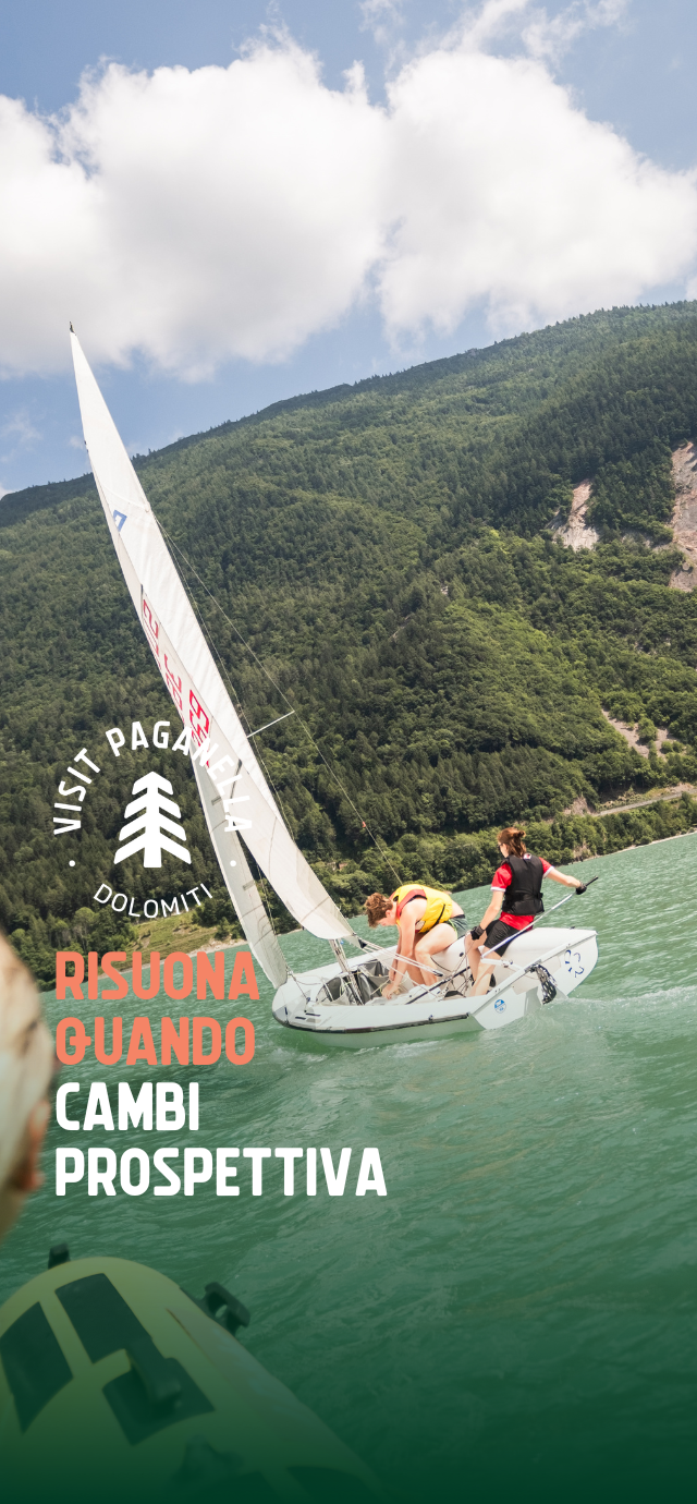 Due persone stanno manovrando una barca a vela su un lago dalle acque verde smeraldo, circondato da un fitto bosco che ricopre le montagne sullo sfondo. Il cielo è parzialmente nuvoloso. La barca è inclinata e in movimento, con la vela tesa dal vento. In primo piano, si intravede un'altra barca. Sulla sinistra dell’immagine compaiono il logo “Visit Paganella” e la scritta “Risuona quando cambi prospettiva”. | © Filippo Frizzera, 2024