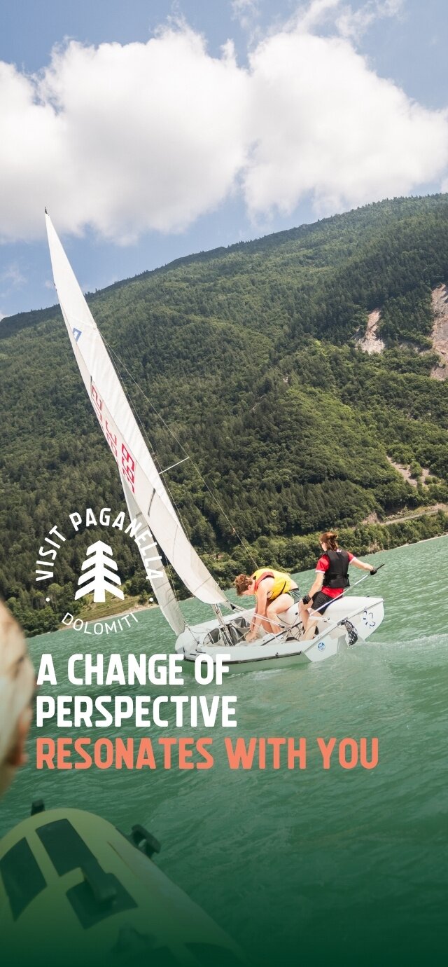 Two people are manoeuvring a sailboat on an emerald-green lake, surrounded by a dense forest covering the mountains in the background. The sky is partly cloudy. The boat is tilted and in motion, with the sail stretched by the wind. In the foreground, another boat can be glimpsed. On the left of the picture are the “Visit Paganella” logo and the inscription “A change of perspective resonates with you”. | © Filippo Frizzera, 2024