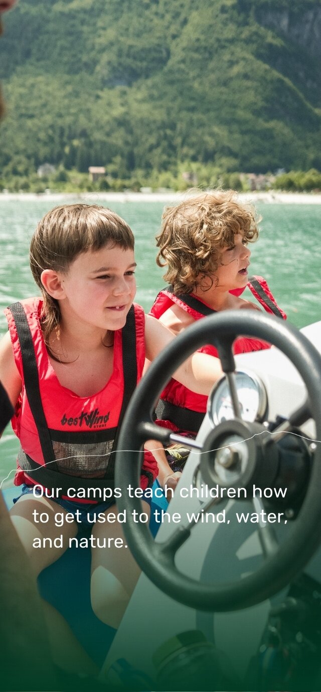  Two children wearing red life jackets are sitting on a sailboat, concentrating and smiling. The rudder can be seen in the foreground. In the background, the green lake is surrounded by wooded mountains. At the bottom, the photo includes a text: ‘Our camps teach children how to get used to the wind, water, and nature.’ | © Filippo Frizzera, 2024